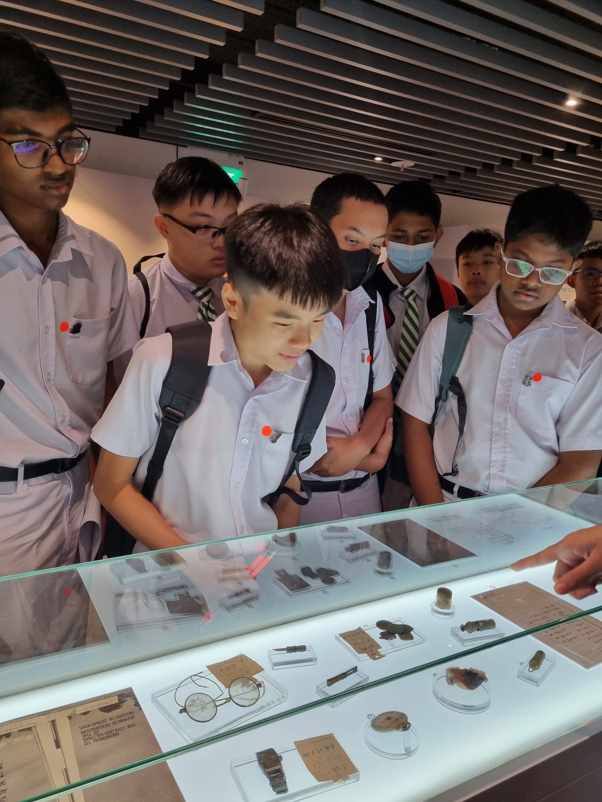 Students view a lit display case of Japanese Occupation-era artefacts, including glasses, a watch, and old documents.